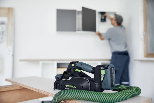 Festool HLC 82 GB Akku-Hobel mit Akkupack und Ladegerät auf grünem Kabel, Anwendung im Hintergrund